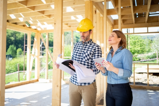 Zwei Personen in einem teilweise gebauten Holzrahmenhaus, eine davon mit gelbem Bauhelm und Bauplänen.